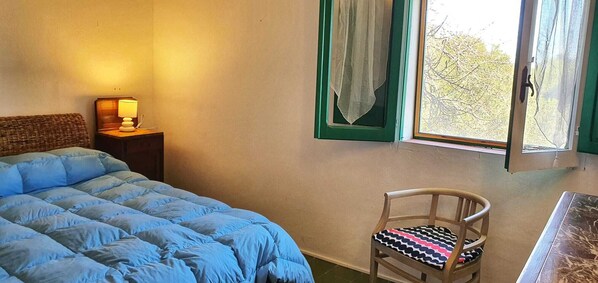 A bedroom with a bed, a chair, a nightstand with a lamp, and a window with curtains.