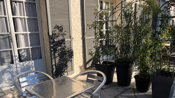 Um pátio com cadeiras de metal e uma mesa de vidro, rodeado por plantas em vasos e um edifício com persianas.