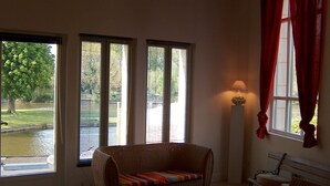 Quarto com sofá de vime, um banco e uma luminária. Três janelas com persianas oferecem vista para um lago e árvores.