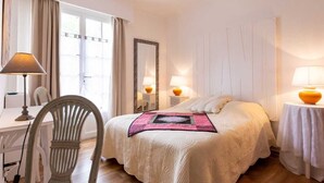 Quarto de hotel com uma cama, duas luminárias de cabeceira, uma cadeira, uma escrivaninha e uma janela com cortinas.