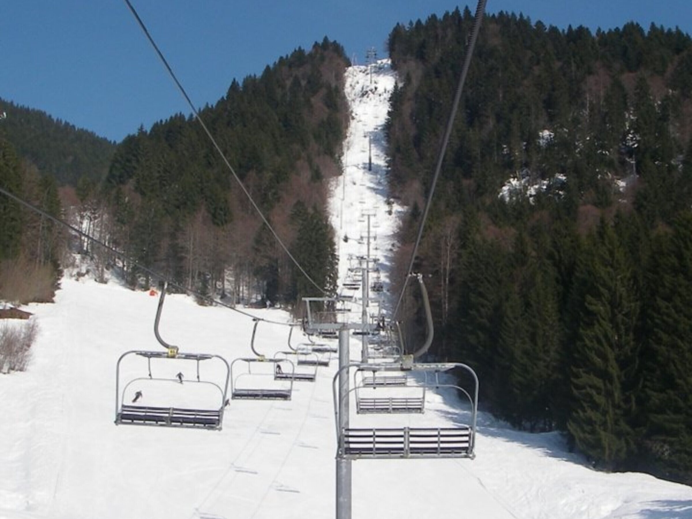 A ski lift with chairs and cables, surrounded by snow-covered slopes and dense forest.