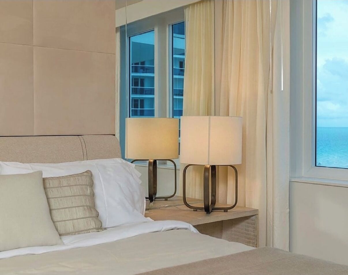 A hotel room with a bed, two bedside lamps, a window with curtains, and a view of the sea.