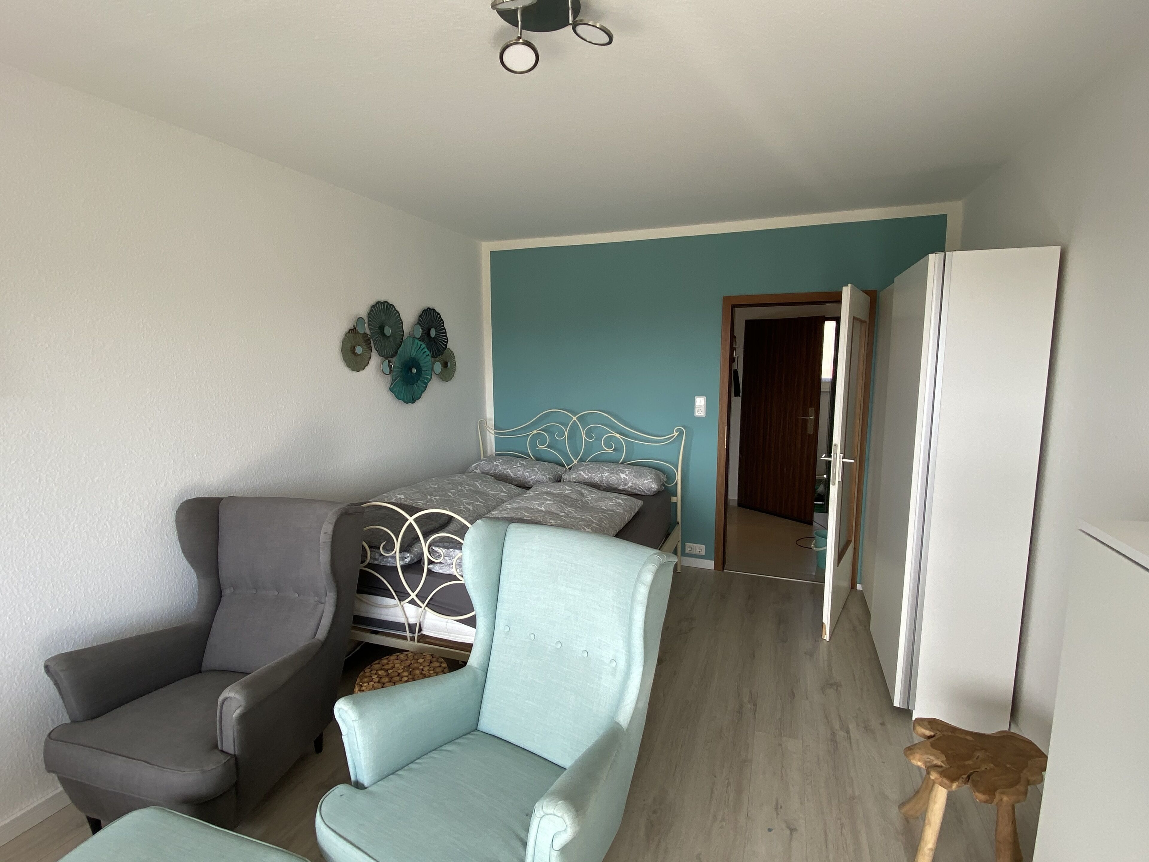 A bedroom with a bed, two armchairs, a wooden stool, a ceiling fan, and a door leading to another room.