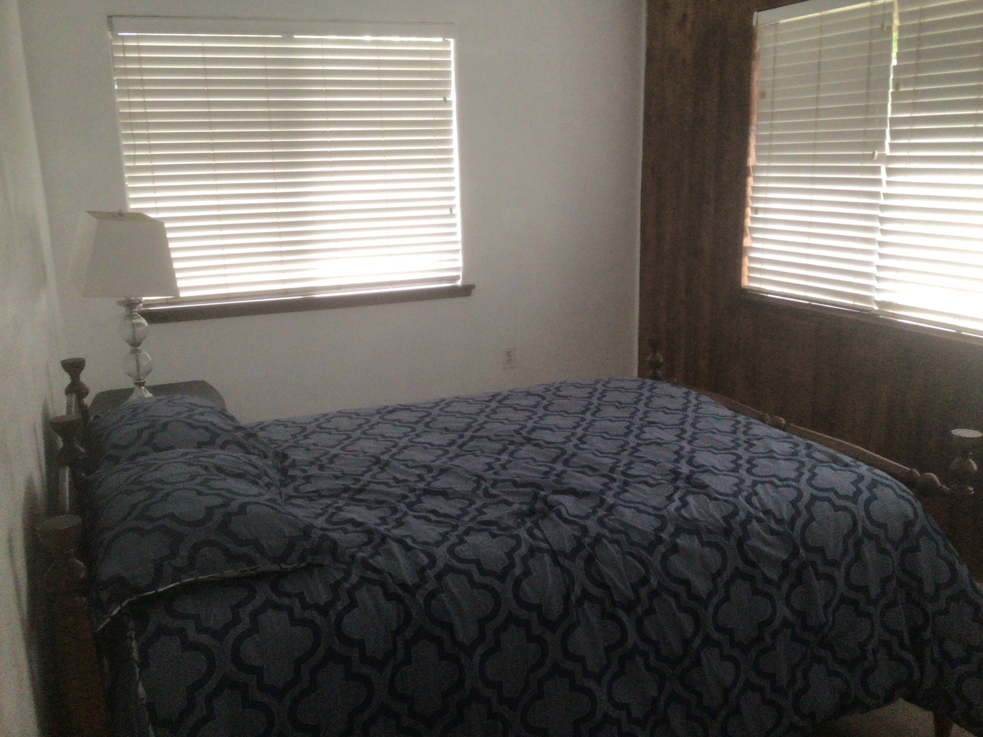 A bedroom with a bed, a nightstand, a lamp, and two windows with blinds.