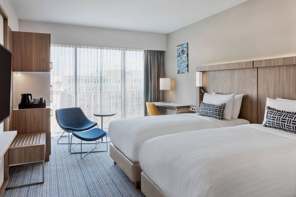 Une chambre d’hôtel avec deux lits, un bureau, une chaise, une télévision et une grande fenêtre avec des rideaux.