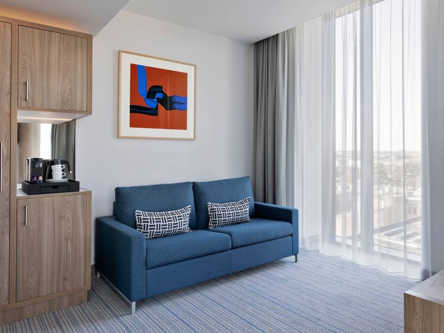 Een moderne hotelkamer met een blauwe bank, een houten kast, een lijst met abstracte kunst, en een groot raam met gordijnen.