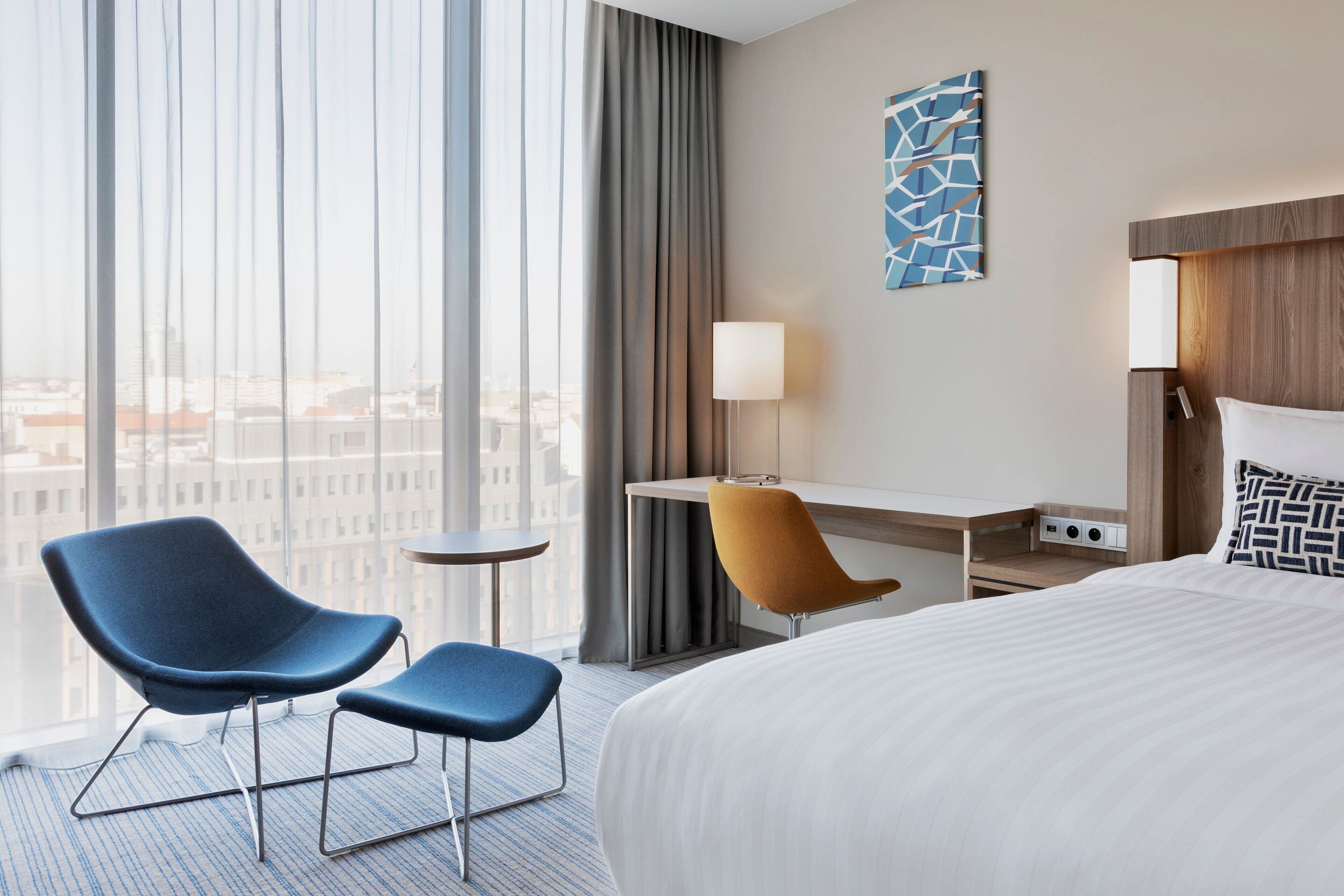 A hotel room with a large bed, two chairs, a desk, a lamp, and a view of the city through large windows.