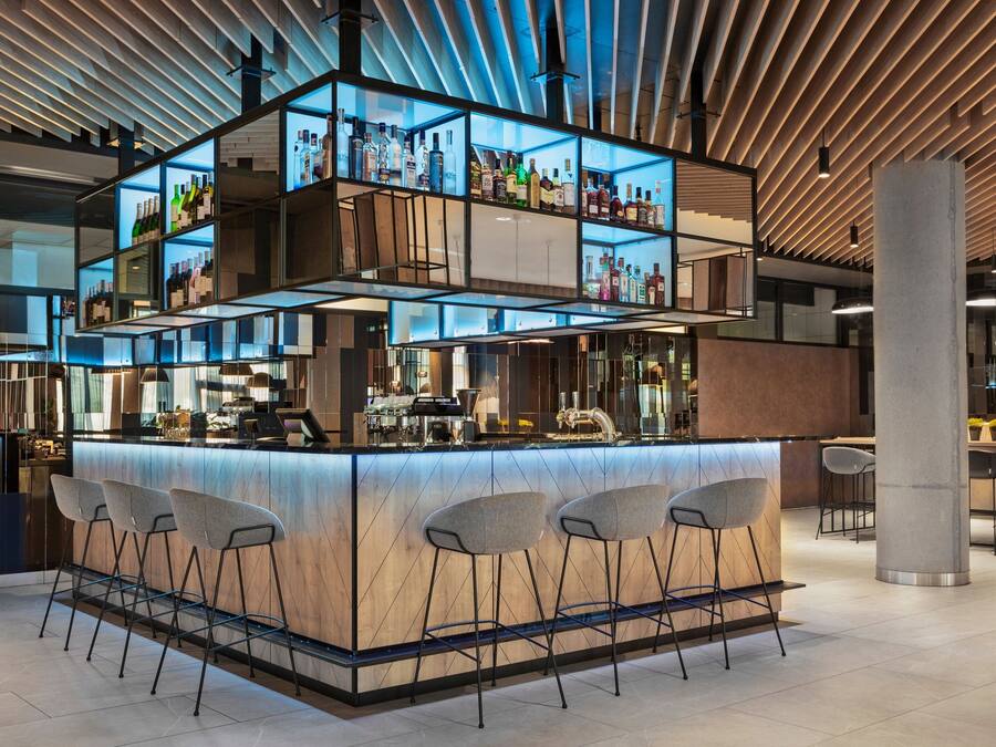 Uma área de bar moderna com um balcão central, diversas prateleiras cheias de garrafas e banquetas.