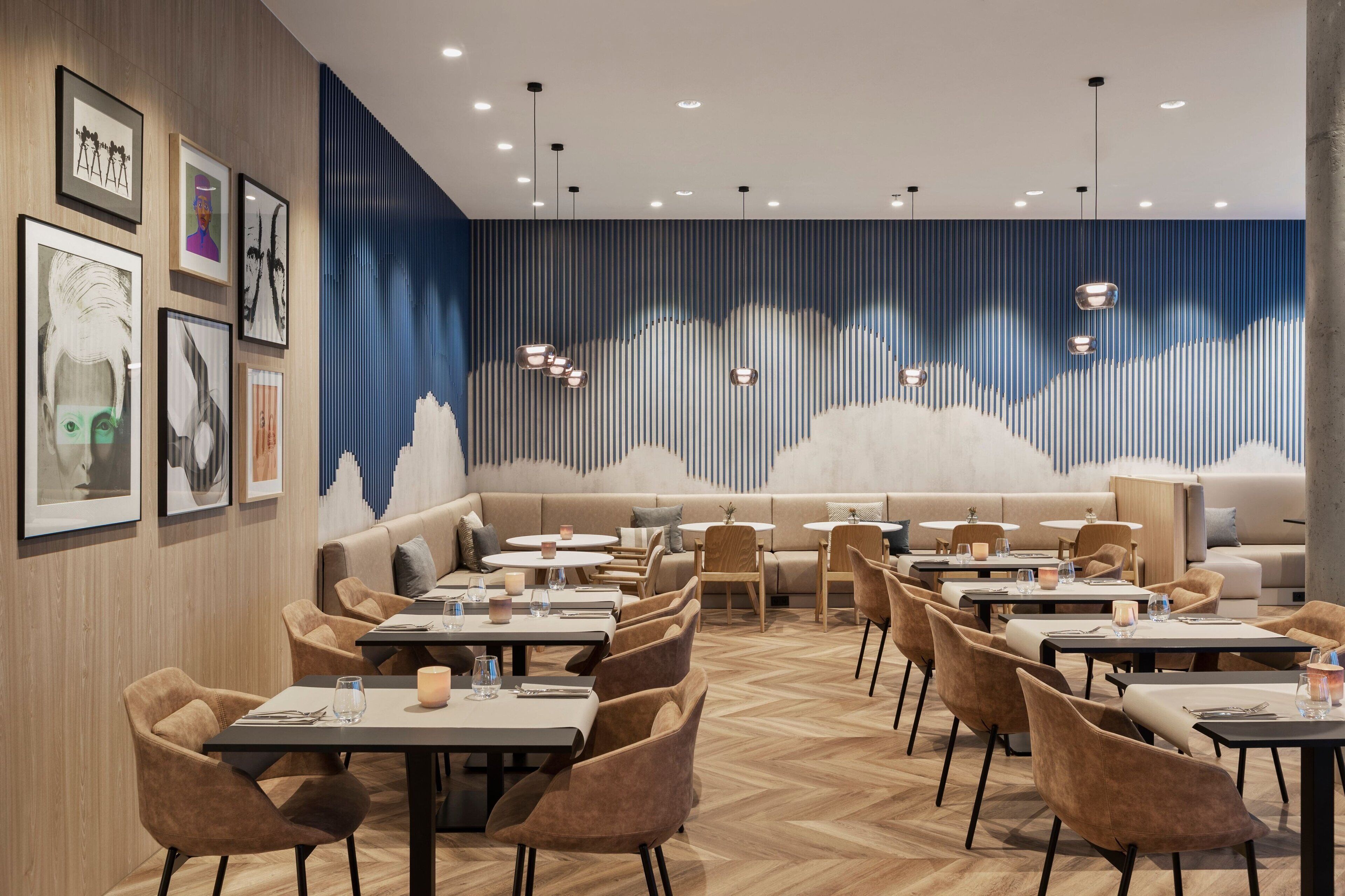A modern restaurant with a wooden herringbone floor, a variety of seating options including tables and a sofa area, and a wall with framed artwork.