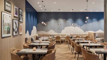 A modern restaurant with a wooden herringbone floor, a variety of seating options including tables and a sofa area, and a wall with framed artwork.
