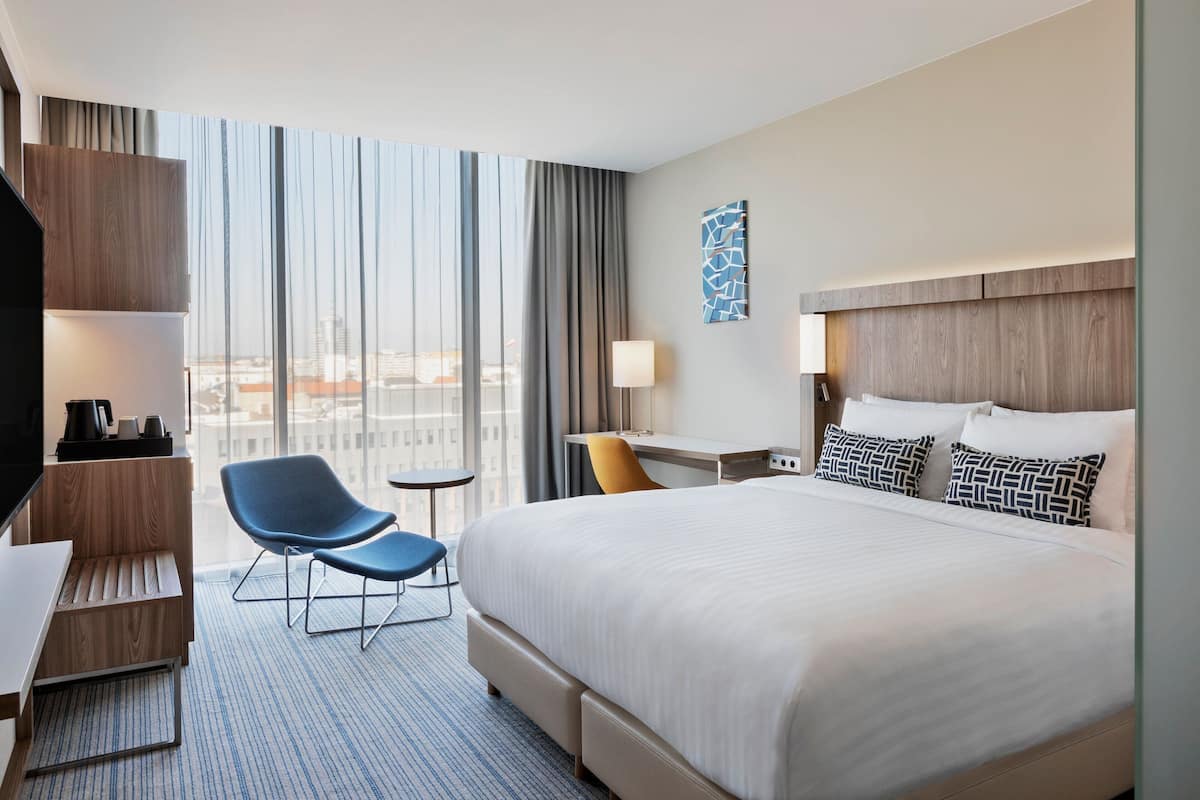 Une chambre d’hôtel avec un grand lit, un bureau, une chaise et une vue sur la ville grâce à de grandes fenêtres.