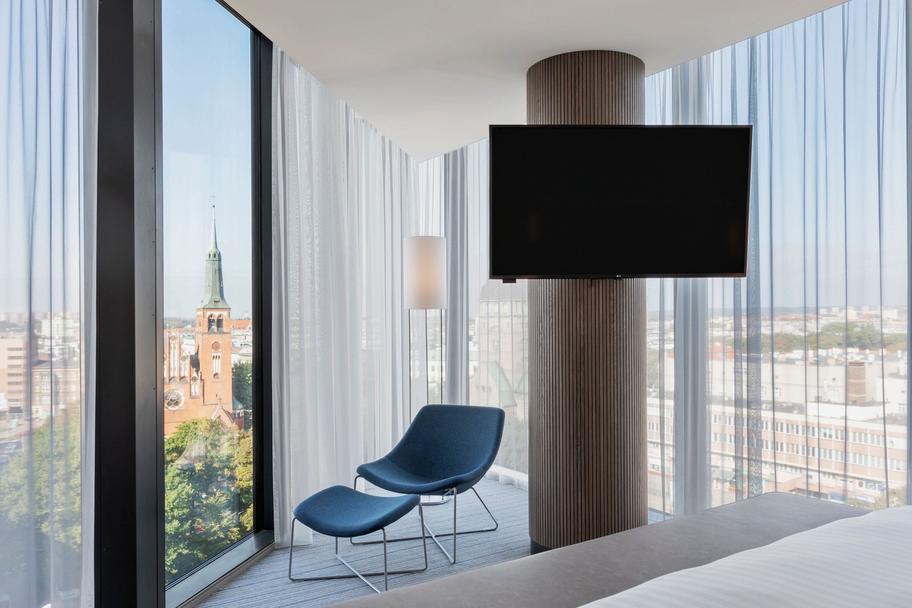Ein modernes Hotelzimmer mit großem Fenster, einem flachbildfernseher an der Wand, einem blauen Sessel und einem Bett.