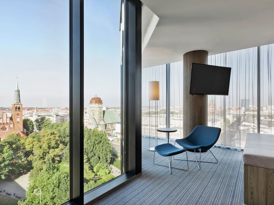 A modern hotel room with a large window offering a city view, a flat-screen TV, a blue chair, and a wooden accent wall.