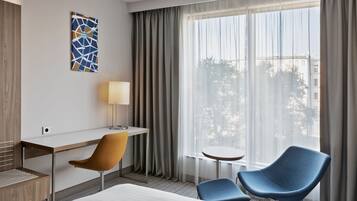 A hotel room with a bed, a desk, two chairs, a lamp, and a large window with curtains.