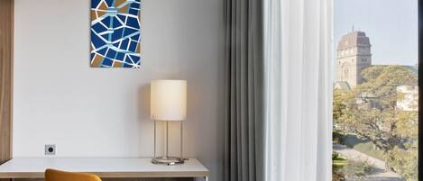 Habitación de hotel con escritorio, una silla, lámpara, ventana con cortinas y vistas a un edificio y árboles.