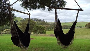 Two hammocks suspended from a wooden structure, set against a backdrop of a grassy field and distant hills.