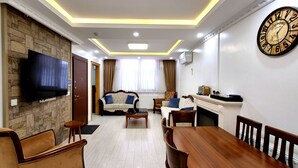 A modern living room with a flat-screen TV, a fireplace, a dining table with chairs, and a wall clock.