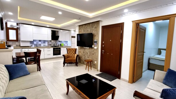 A modern living room with a sofa, coffee table, and a TV. There is a kitchen in the background with white cabinets and a stone accent wall.