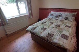 A bedroom with a wooden floor, a radiator, a window with curtains, and a bed with a quilt.