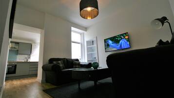 A living room with a sofa, a coffee table, a TV mounted on the wall, and a kitchen in the background.
