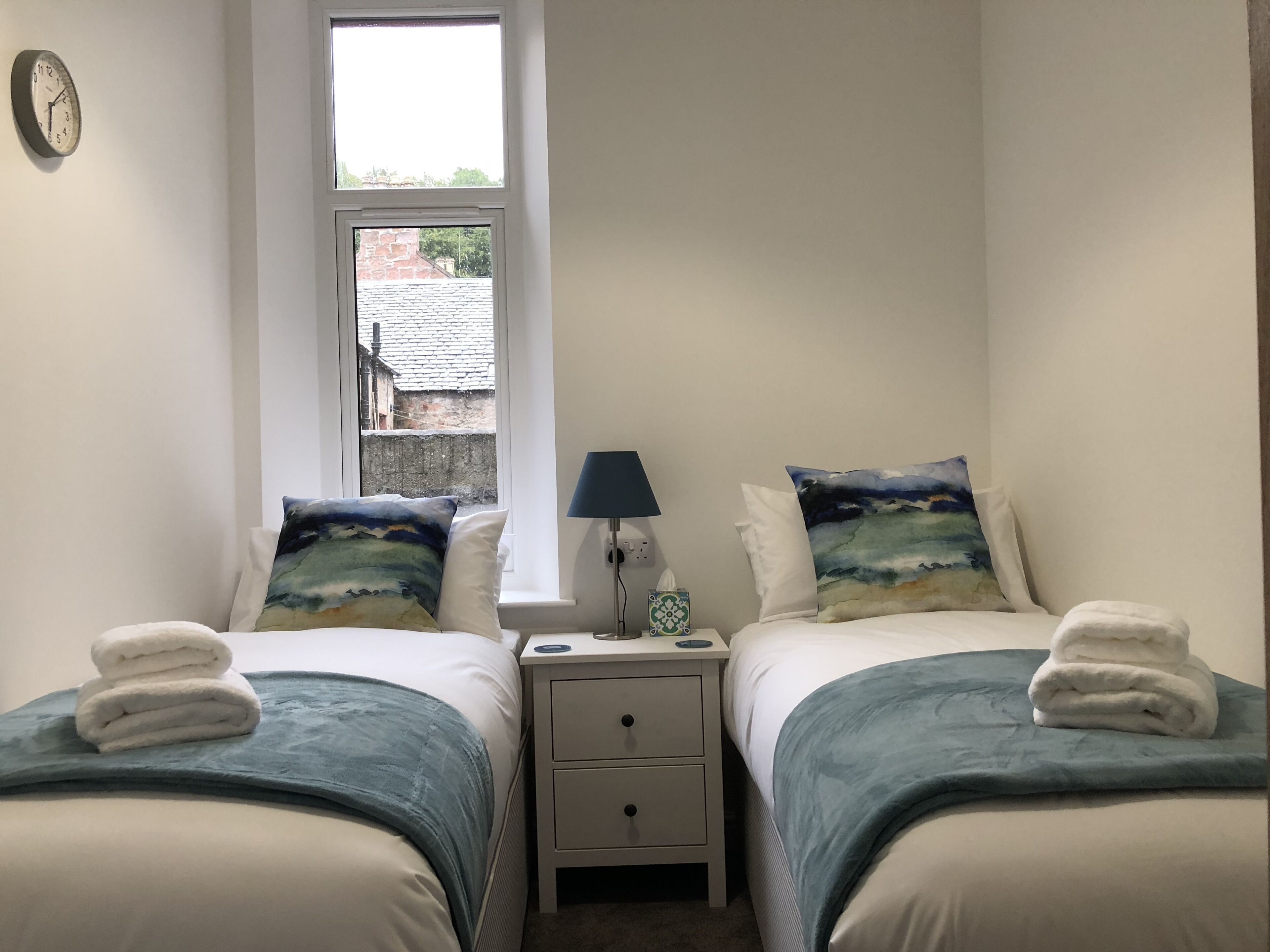 A bedroom with two beds, each with a blue blanket and a pillow with a landscape design. There is a bedside table with a lamp and a tissue box. A clock is visible on the wall.