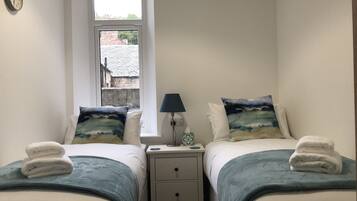 A bedroom with two beds, each with a blue blanket and a pillow with a landscape design. There is a bedside table with a lamp and a tissue box. A clock is visible on the wall.