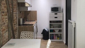 Cocina compacta con microondas, un refrigerador pequeño y un mueble blanco. En primer plano, una mesa de comedor con una silla.