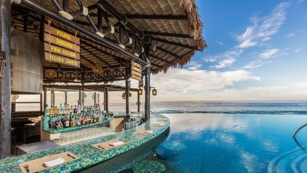 An infinity pool with a bar area featuring a tropical thatched roof and a clear view of the ocean.