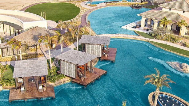 Vista aérea de un resort con una gran piscina central, bungalows sobre el agua y una zona ajardinada con palmeras.