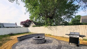Un jardin avec un foyer en pierre, un barbecue et une clôture blanche.