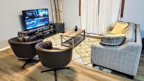 Un salon équipé d’un téléviseur à écran plat, de deux fauteuils en cuir, d’un canapé gris agrémenté d’un coussin à rayures, d’une table basse en bois et d’un rideau blanc.