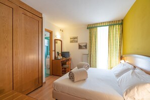 A bedroom with a bed, a wooden wardrobe, a desk with a computer, a mirror, and a window with curtains.