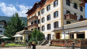 A multi-story hotel with balconies, a patio area with outdoor seating, and a mountain backdrop. - Hotel Posta (Aprica)