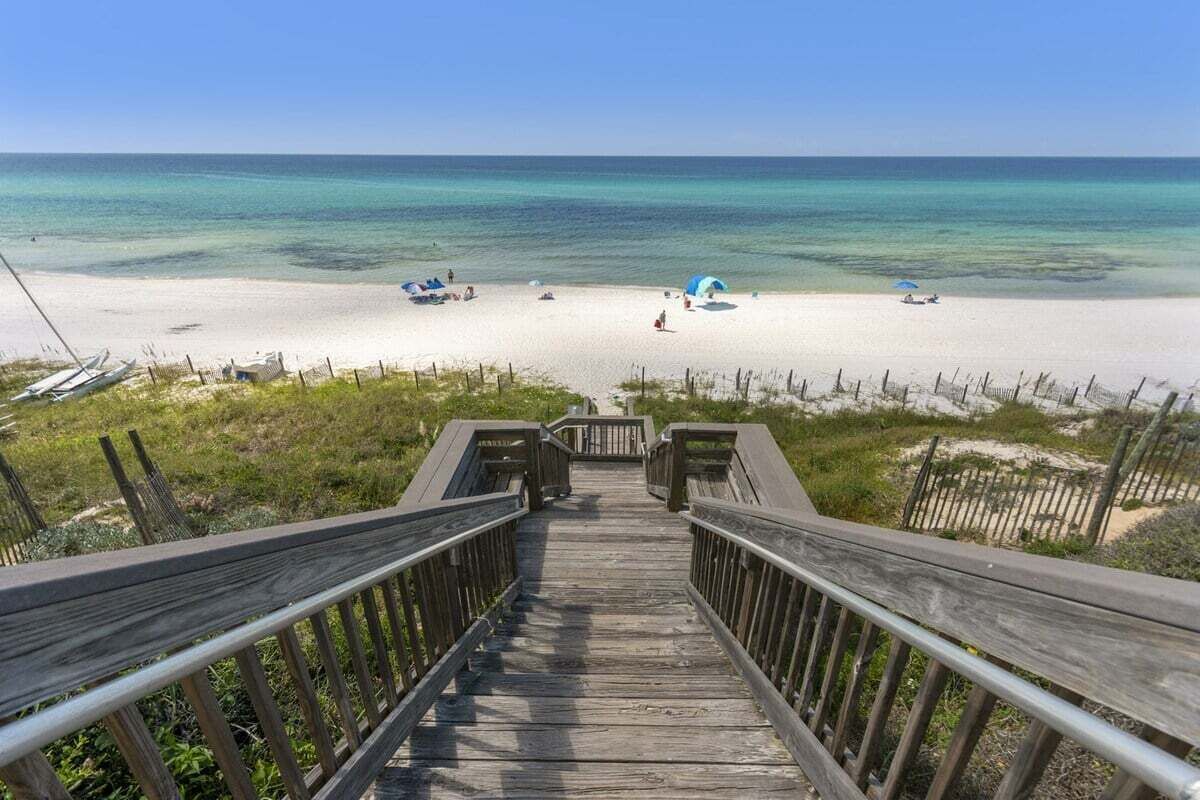 Une promenade en bois qui mène à une plage où l’eau est d’un bleu turquoise limpide et le sable est blanc.