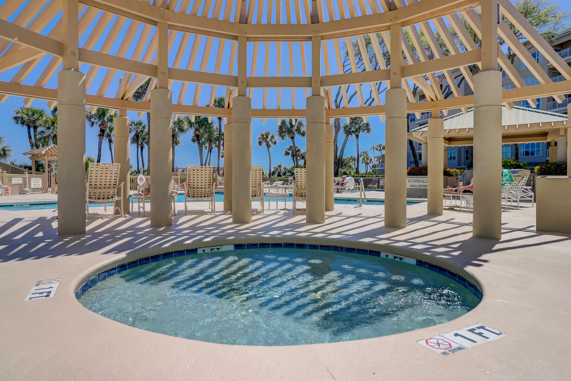 Una piscina circolare con una pergola sopra, circondata da poltrone prendisole e palme.