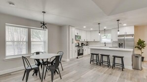 A modern kitchen with white cabinets, a central island with stools, and a dining area with a round table and chairs. - Gorgeous Sunny Home for Big Groups w/ Private Pool (Garland)