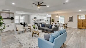 A spacious living area with a sofa, coffee table, and dining table. A kitchen with white cabinets and a ceiling fan is visible in the background. - Gorgeous Sunny Home for Big Groups w/ Private Pool (Garland)