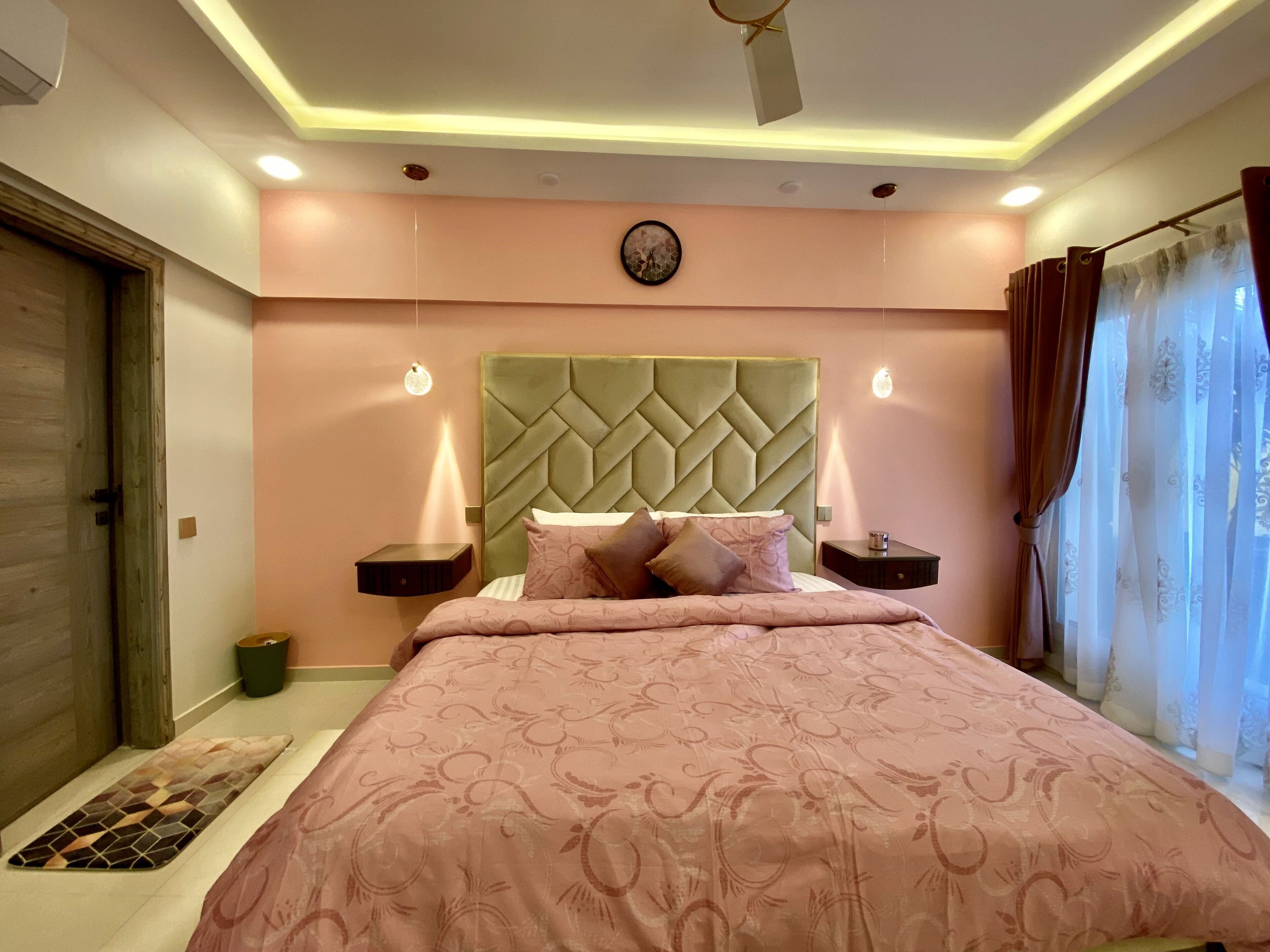 A bedroom with a large bed, a headboard with a diamond pattern, bedside tables with lamps, a clock on the wall, and a window with curtains.