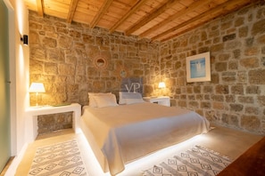 A bedroom with a stone wall, a bed with white bedding, two bedside lamps, and a framed picture on the wall. - Villa Nirvana- Calm, Relaxing and no Wind! (Mykonos)