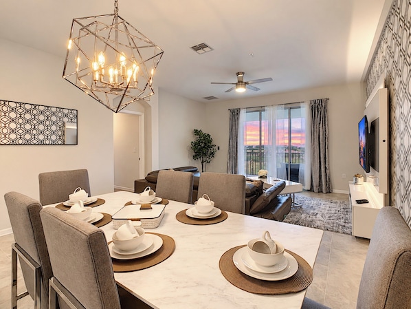A modern dining area with a geometric chandelier, a ceiling fan, and a view of a sunset through large windows.