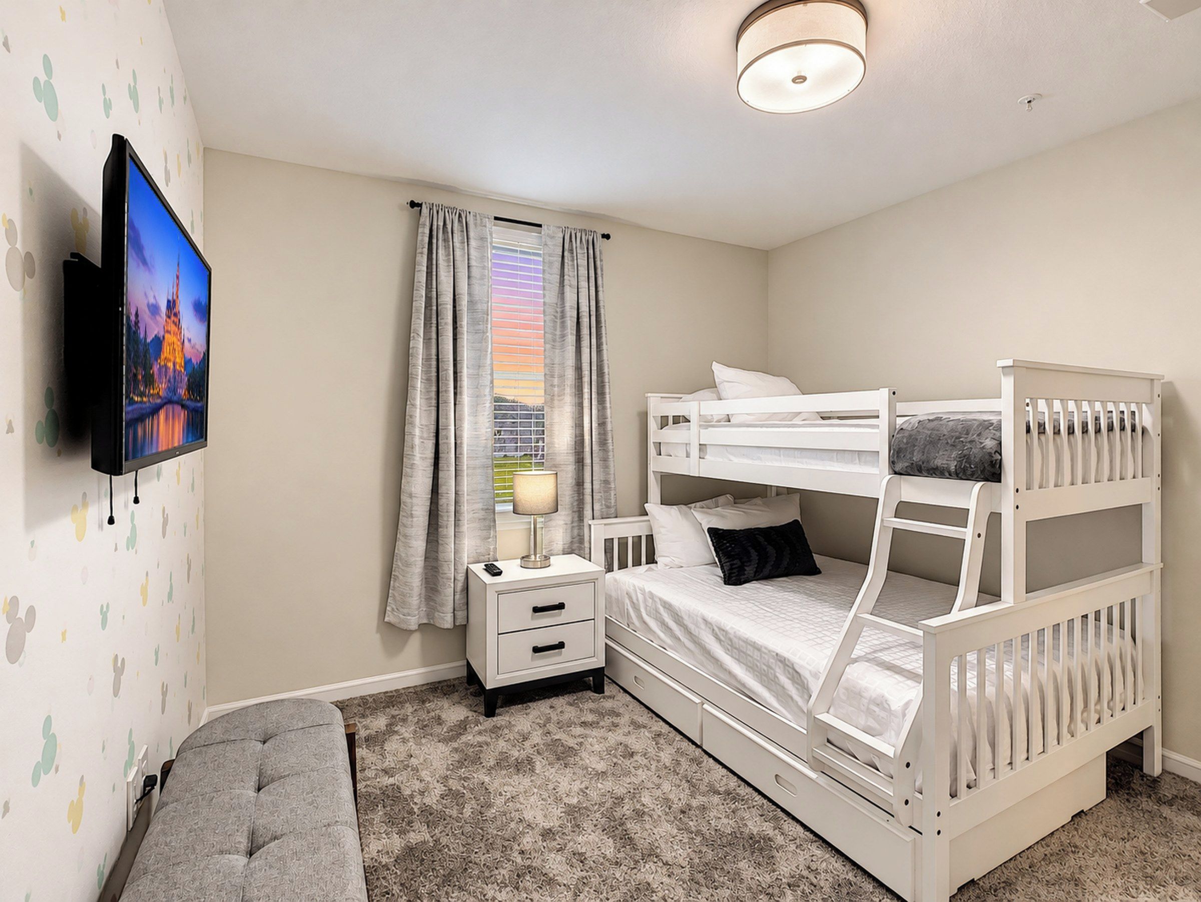 A bedroom with a bunk bed, a bedside table with a lamp, a TV mounted on the wall, and a window with curtains.