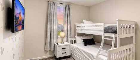 A bedroom with a bunk bed, a bedside table with a lamp, a TV mounted on the wall, and a window with curtains.
