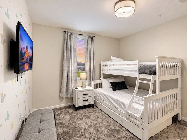 A bedroom with a bunk bed, a bedside table with a lamp, a TV mounted on the wall, and a window with curtains.