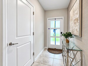 A hallway with a white door, a glass entry door with frosted glass panels, a doormat, and a glass console table with a plant.