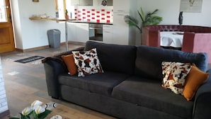 A living room with a dark grey sofa, two patterned throw pillows, a small table with a white tablecloth, and a kitchen in the background with white cabinets and red tiles. - Pleasant country cottage in a quiet location (Wirwignes)