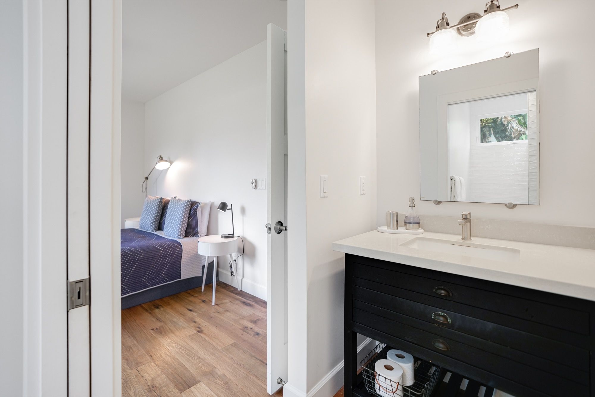 A bedroom with a bed, bedside table, lamp, and a bathroom with a sink, mirror, and toilet paper storage.