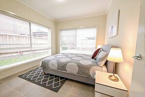A bedroom with a bed, bedside lamp, and a window with a view of a fence. - Point Cook 6 BRM 4-Bathroom House for Big Group (Point Cook)