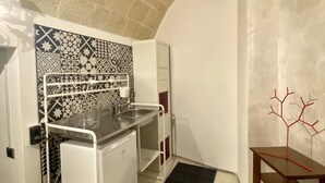 A compact kitchen with a patterned backsplash, a white refrigerator, and a small table with a red decorative piece. - Abitazione Tipica (Lecce)