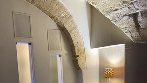 A bedroom with a bed, two pillows, a nightstand, a lamp, and a stone archway. - Abitazione Tipica (Lecce)