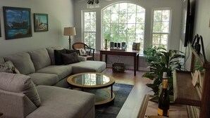 A living room with a grey sectional sofa, a round coffee table, and a bookshelf. There are large windows with a view of trees, a lamp, and a television. - Beautiful, 3 br/2 bath condo in Pawleys Island, SC. Minutes from the beach. (Pawleys Island)